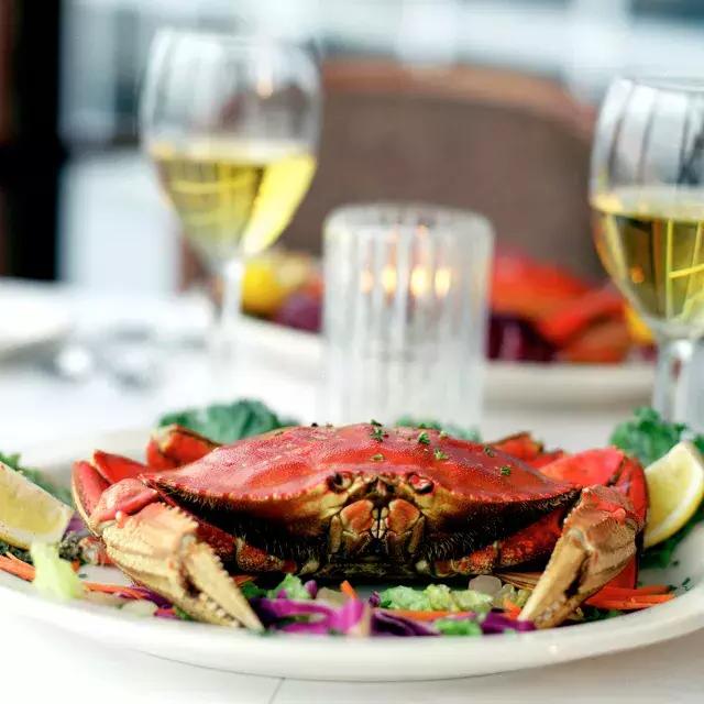 A Dungeness crab sits on a plate in a restaurant with two glasses of white wine 在 background.